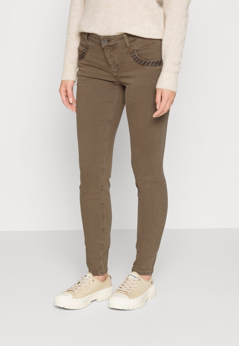 Jeans skinnies marrones con bolsillos decorativos, hechos de una mezcla de algodón. Combinados con zapatillas beiges con suelas texturizadas.