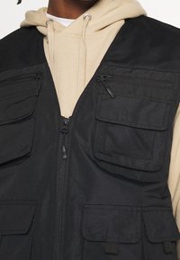 Sudadera beige con cordones, llevada debajo de un chaleco negro que presenta múltiples bolsillos con cremallera y solapa.