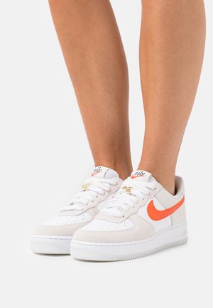 Buty Nike Air Force 1 z białą skórzaną cholewą z pomarańczowymi detalami, zamszowymi nakładkami i teksturowaną gumową podeszwą.