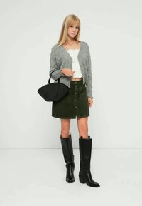 Siv pleteni kardigan, beli kratki top, olivno zelena krilo z gumbi spredaj in črni škornji do kolen. Črna torbica s ukrivljenim dizajnom.