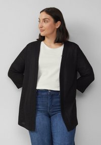 Zwarte gebreide cardigan met een open voorkant, driekwart mouwen en een relaxte fit, gecombineerd met een wit t-shirt en blauwe jeans.