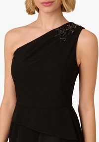 Czarna sukienka na jedno ramię z gładkiego materiału charakteryzuje się drapowaną konstrukcją, falbaniastym dołem oraz zdobieniem z koralików na ramieniu.