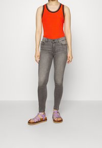 Top de tirantes rojo con canalé y ribete negro, vaqueros ajustados grises y sandalias moradas con tiras y suela marrón. La modelo está de pie contra un fondo liso.