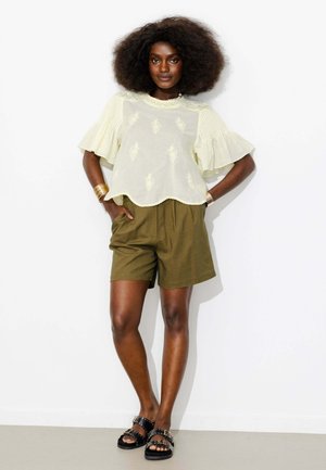 Femme avec une coiffure afro naturelle porte un chemisier crème brodé avec des manches à volants, un short kaki et des sandales noires cloutées.