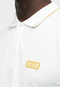 Κοντινή λήψη ενός λευκού πόλο μπλουζιού με γιακά με χρυσές λεπτομέρειες και χρυσή ετικέτα με το λογότυπο "Versace Jeans Couture" στο στήθος.