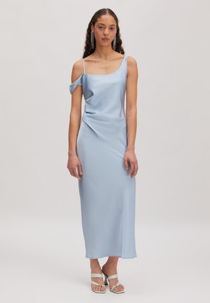 Frau, die ein hellblaues Satin-Midikleid mit asymmetrischen Trägern und weißen Sandaletten mit Absatz trägt, steht vor einem einfarbigen Hintergrund.