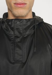 Svart huvajacka av glänsande, lätt material. Har en framficka, dragsnören och en tryckknappslåsning med logotypdetalj.