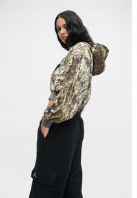 Φούτερ με καμουφλάζ σε γήινα χρώματα, διαθέτει κουκούλα και ριμπ μανσέτες. Συνδυάζεται με μαύρα τζόγκινγκ που επιδεικνύουν λογότυπο στην τσέπη.