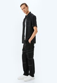 Camisa negra de manga corta con costuras contrastantes en blanco y dos bolsillos frontales, combinada con pantalones cargo negros y zapatillas blancas.