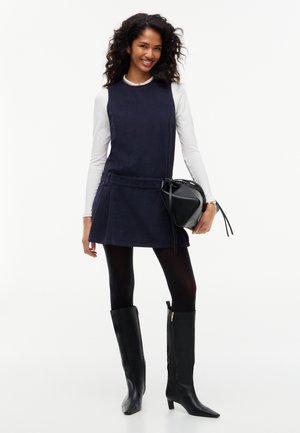 Femme portant une robe sans manches bleu marine par-dessus un haut blanc à manches longues, des collants noirs, des bottes noires jusqu'au genou et tenant un sac à main noir.