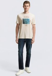 T-shirt corta in cotone color crema con stampa grafica al centro, con design a onde blu e elementi di testo. Abbinata a jeans blu scuro.