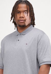 Polo shirt grigio a maniche corte con colletto, caratterizzato da un piccolo logo con accenti rossi, bianchi e blu sul petto, realizzato in tessuto morbido.