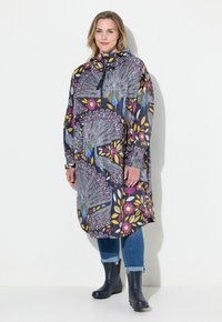 Manteau long à capuche ornée d'un motif florale vibrant et de plumes de paon dans des teintes de violet, jaune et bleu, associé à un jean en denim et des bottes noires.