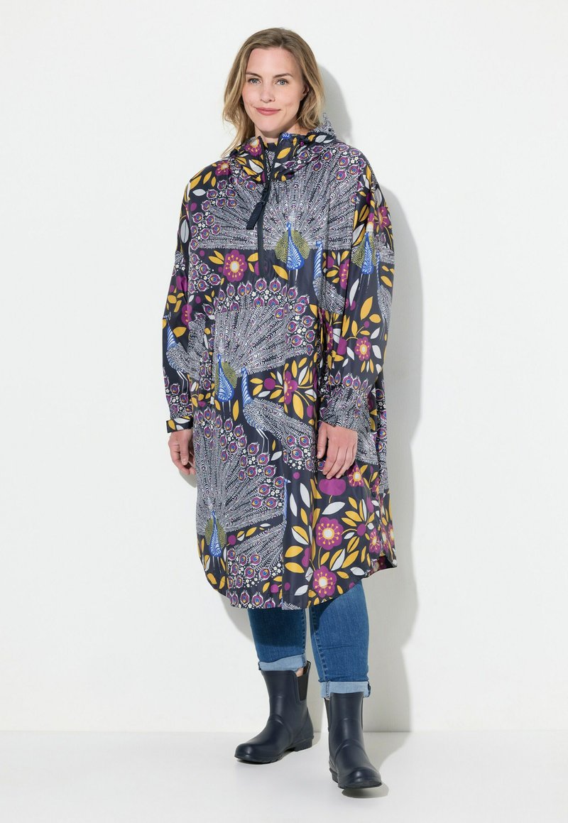 Manteau long à capuche ornée d'un motif florale vibrant et de plumes de paon dans des teintes de violet, jaune et bleu, associé à un jean en denim et des bottes noires.