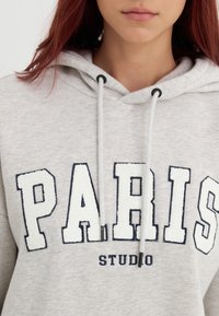 Svetlo šedá mikina s kapucňou s bielym chĺpkovým nápisom "PARÍŽ" a "ŠTÚDIO" pod ním. Obsahuje kapucňu na šnúrku a textúrovanú látku.