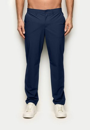 Mężczyzna w granatowych prostych spodniach i białych trampkach, stoi na tle jasnego, gładkiego tła.