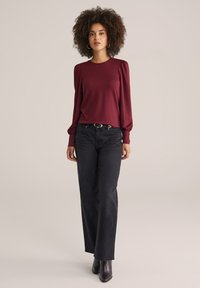 Haut texturé bordeaux à manches longues avec épaulettes bouffantes, associé à un jean noir taille haute et des bottes noires jusqu'aux genoux.