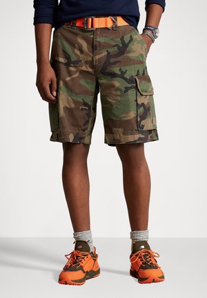 Personne portant un short camouflage avec plusieurs poches, une ceinture orange, des chaussettes grises et des chaussures de sport orange et marron.
