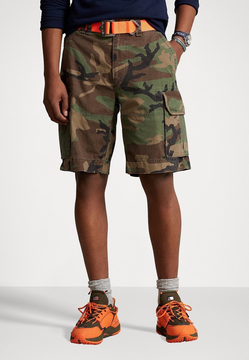 Personne portant un short camouflage avec plusieurs poches, une ceinture orange, des chaussettes grises et des chaussures de sport orange et marron.