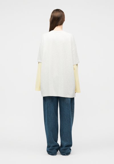 Άτομο με μακριά καστανά μαλλιά, που φοράει μια φαρδιά λευκή μπλούζα πάνω από μακριά μπεζ μανίκια και χαλαρά μπλε τζιν, όρθιο πλάτη προς τα εμπρός.