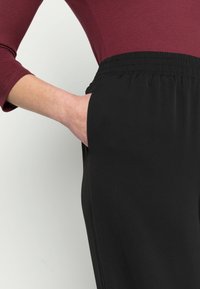 Černé kalhoty s elastickým pasem a hladkou strukturou, kombinované s bordó přiléhavým topem. Viditelné detaily kapes na boku.