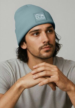 Light blue ribbed knit beanie with a rectangular patch featuring a camera icon. Model wears a gray t-shirt, hands clasped in front.