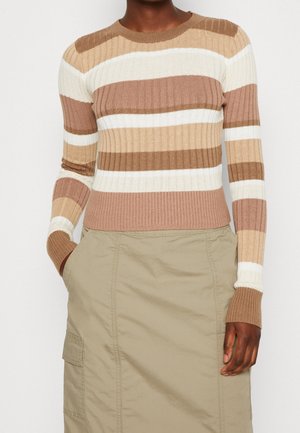 Persona que lleva un suéter de canalé con rayas horizontales beige, marrón y blancas, combinado con una falda caqui con bolsillos y la mano en el bolsillo.