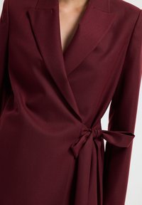 Donna che indossa un blazer dress tailored di un profondo borgogna con un fiocco annodato di lato e rever affilati.