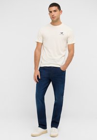 Camiseta de algodón beige con un gráfico impreso, combinada con jeans de mezclilla azul oscuro y zapatillas blancas. Corte clásico y diseño simple.