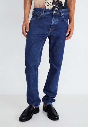 Vaqueros de denim azul con un corte recto, que presentan un diseño clásico de cinco bolsillos y detalles de costura visibles. Llevados con zapatos negros.