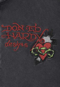 Tmavá látka s červeným vyšitým textem "DON ED HARDY designs" vedle červeného srdce propíchnutého černobílou dýkou, která je obtočena zelenými vinnými listy.