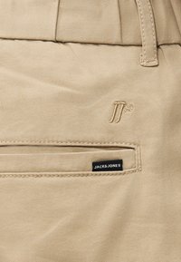 Μπεζ chinos με πίσω τσέπη που έχει μια μαύρη ετικέτα "JACK & JONES" και ένα μικρό κεντημένο λογότυπο. Ομαλό ύφασμα με λεπτομερείς ραφές.