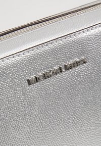 Funda de cuero texturizado plateado con una cremallera visible y el nombre de la marca "MICHAEL KORS" en relieve en la parte frontal.