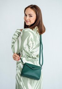 Zelená kožená kabelka ve tvaru obálky, s klopovým zavíráním a dlouhým, flexibilním popruhem. Jemné prošívání zvýrazňuje design.