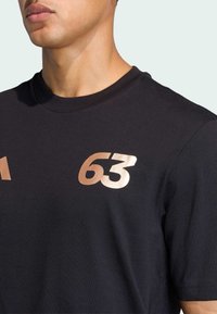 Musta t-paita, jossa on pyöreä pääntie ja lyhyet hihat. Edessä on metallinhohtoinen ruusukultainen "63" ja "1" -printti.