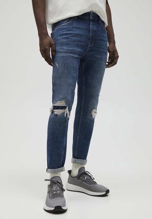 Hombre vistiendo jeans azules desgastados con los dobladillos subidos, calcetines blancos, zapatillas grises y una camiseta blanca frente a un fondo claro y liso.