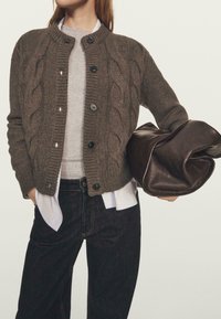 Bruine kabelgebreide cardigan met een ronde halslijn, voorzien van zwarte knopen. Gedragen over een grijze blouse en vergezeld van een donkerbruine tas.