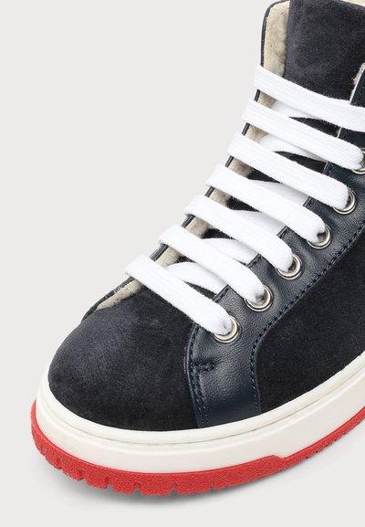 Gros plan d’une basket montante noire et marine avec des lacets blancs, une semelle blanche et une semelle rouge, montrant des textures en suède et cuir.