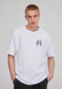 T-shirt bianco in cotone a maniche corte, con un design grafico nero nell'area sinistra del petto. Vestibilità rilassata e scollo rotondo.