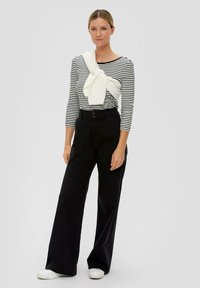 Gestreiftes schwarz-weißes Langarmshirt kombiniert mit weiten schwarzen Hosen. Ein weißer Pullover ist lässig um die Schultern gebunden. Weiße Schuhe.