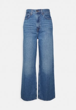 Jean en denim bleu taille haute avec jambes larges, fermeture avant par boutons et fermeture éclair, et poches avant et arrière, présenté sur un fond uni.