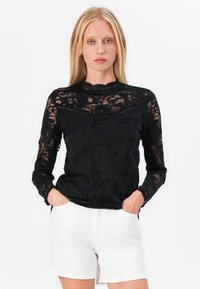 Black lace long-sleeve top with floral pattern, high neck, and scalloped detailing, paired with white shorts.