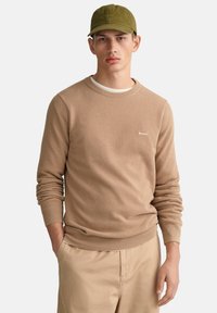 Beige Strickpullover mit strukturiertem Muster und Rundhalsausschnitt, kombiniert mit einer olivgrünen Mütze und hellbeigen Hosen.