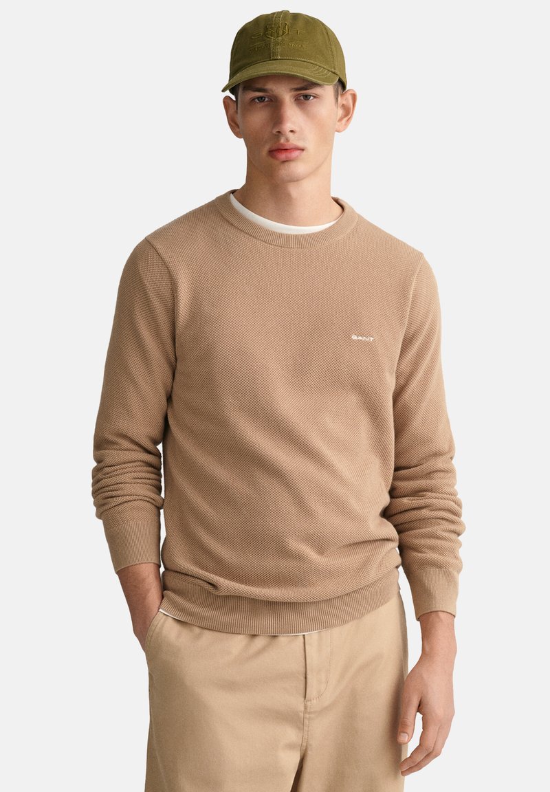 Beige Strickpullover mit strukturiertem Muster und Rundhalsausschnitt, kombiniert mit einer olivgrünen Mütze und hellbeigen Hosen.