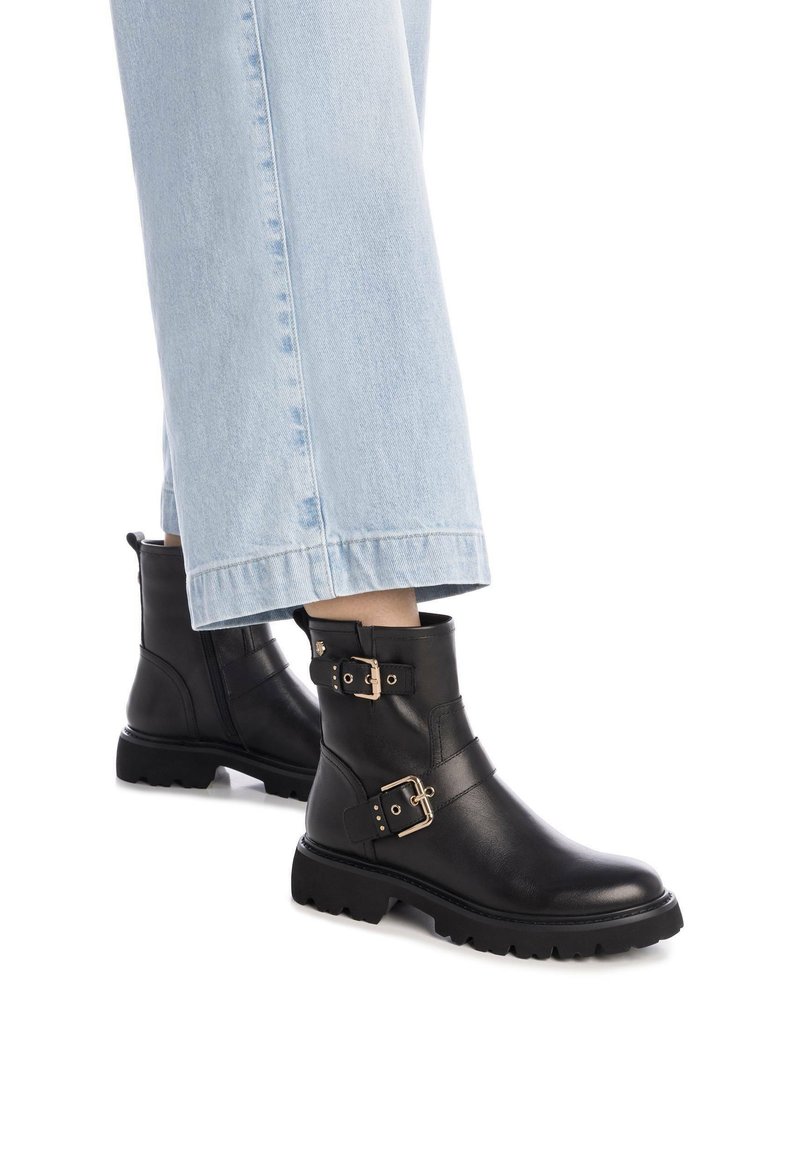 Botas de tobillo de cuero negro con acentos de doble hebilla y suelas de goma resistentes, combinadas con pantalones de mezclilla recortados claros.