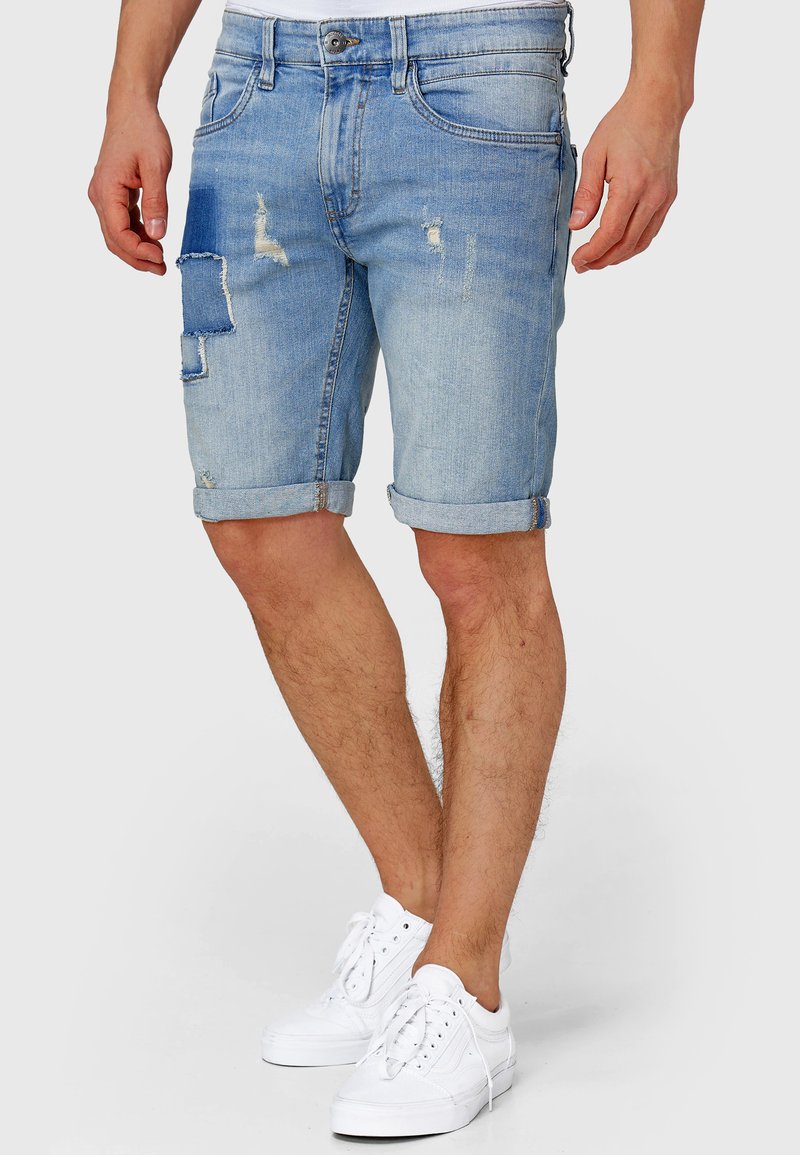 Pantaloni scurți din denim albastru deschis cu design deteriorat cu patchwork, tivuri rulate, stil de cinci buzunare și finisaj decolorat.