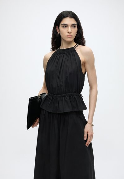 Γυναίκα με μακριά σκούρα μαλλιά που φοράει αμάνικη μαύρη μπλούζα με halter και φαρδιά μαύρη παντελόνα, κρατώντας μαύρο φάκελο σε απλό φόντο.