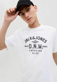 Balta medvilninė marškinėliai su juodu grafiniu tekstu: "JACK & JONES PREMIUM D.N.M. PREKYBA SUPERIOR BLUE." Modelis dėvi juodą kepurę.