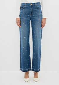 Niebieskie dżinsy z szerokimi nogawkami, zdobione haftowanymi perłami z przodu oraz frędzlami na dole. Zawierają klasyczny pięcio-kieszeniowy design oraz zapięcie na guzik.