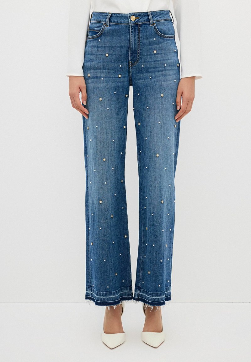 Niebieskie dżinsy z szerokimi nogawkami, zdobione haftowanymi perłami z przodu oraz frędzlami na dole. Zawierają klasyczny pięcio-kieszeniowy design oraz zapięcie na guzik.